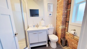 A bathroom with a white vanity, a toilet, a glass shower enclosure, a brick accent wall, and a window with blinds. - Craft District Pet-Friendly Condo by Heirloom (Duluth)