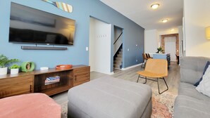 A modern living room with a flat-screen TV, wooden entertainment unit, and comfortable seating. A staircase leads to a kitchen area. - Craft District Pet-Friendly Condo by Heirloom (Duluth)