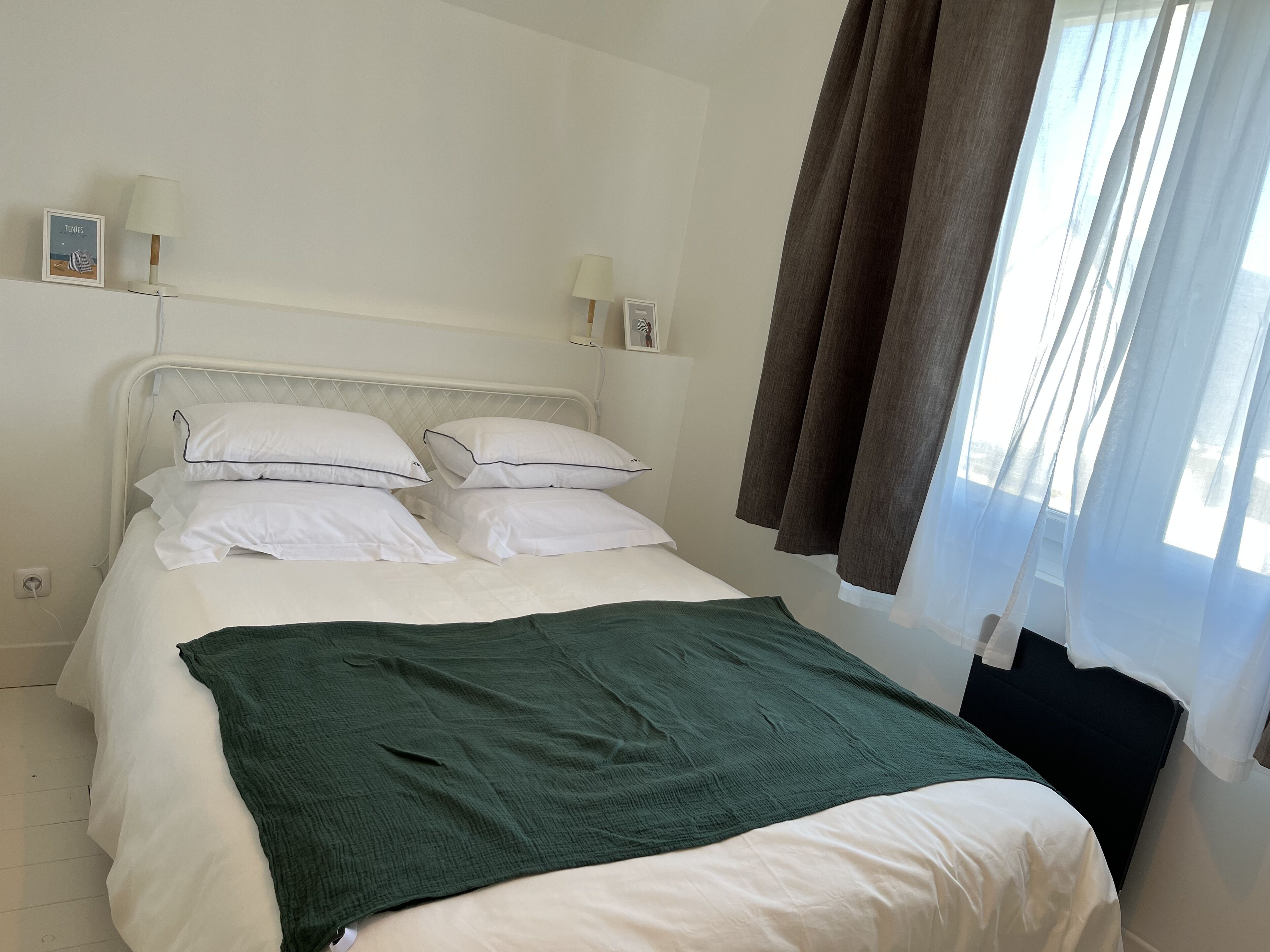 Un lit bien fait, avec du linge de lit blanc et un plaid vert, une lampe de chevet et un tableau encadré au mur.