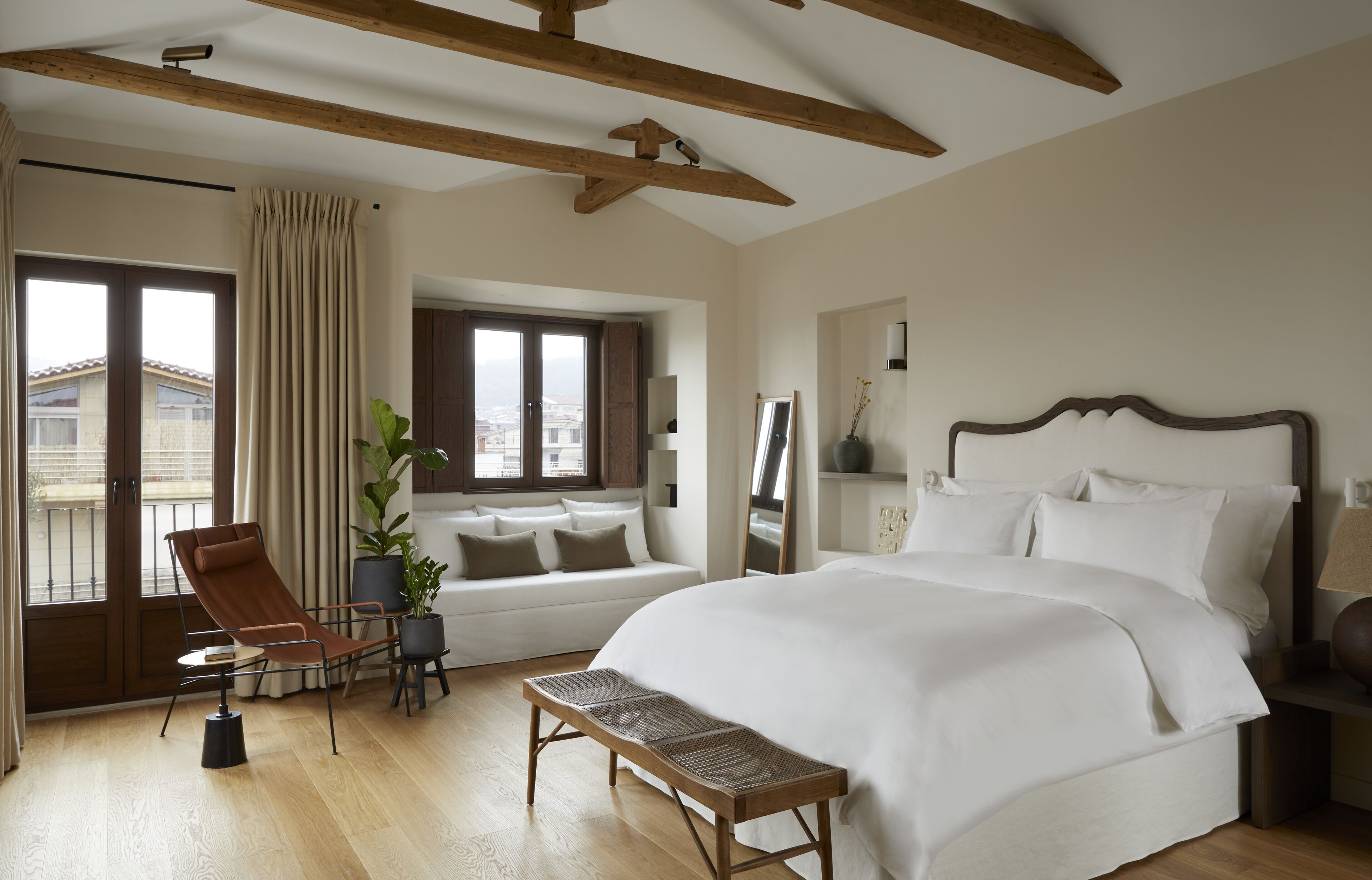 Quarto com uma cama grande, uma cadeira, um banco, uma planta em vaso e vista da cidade pela janela.