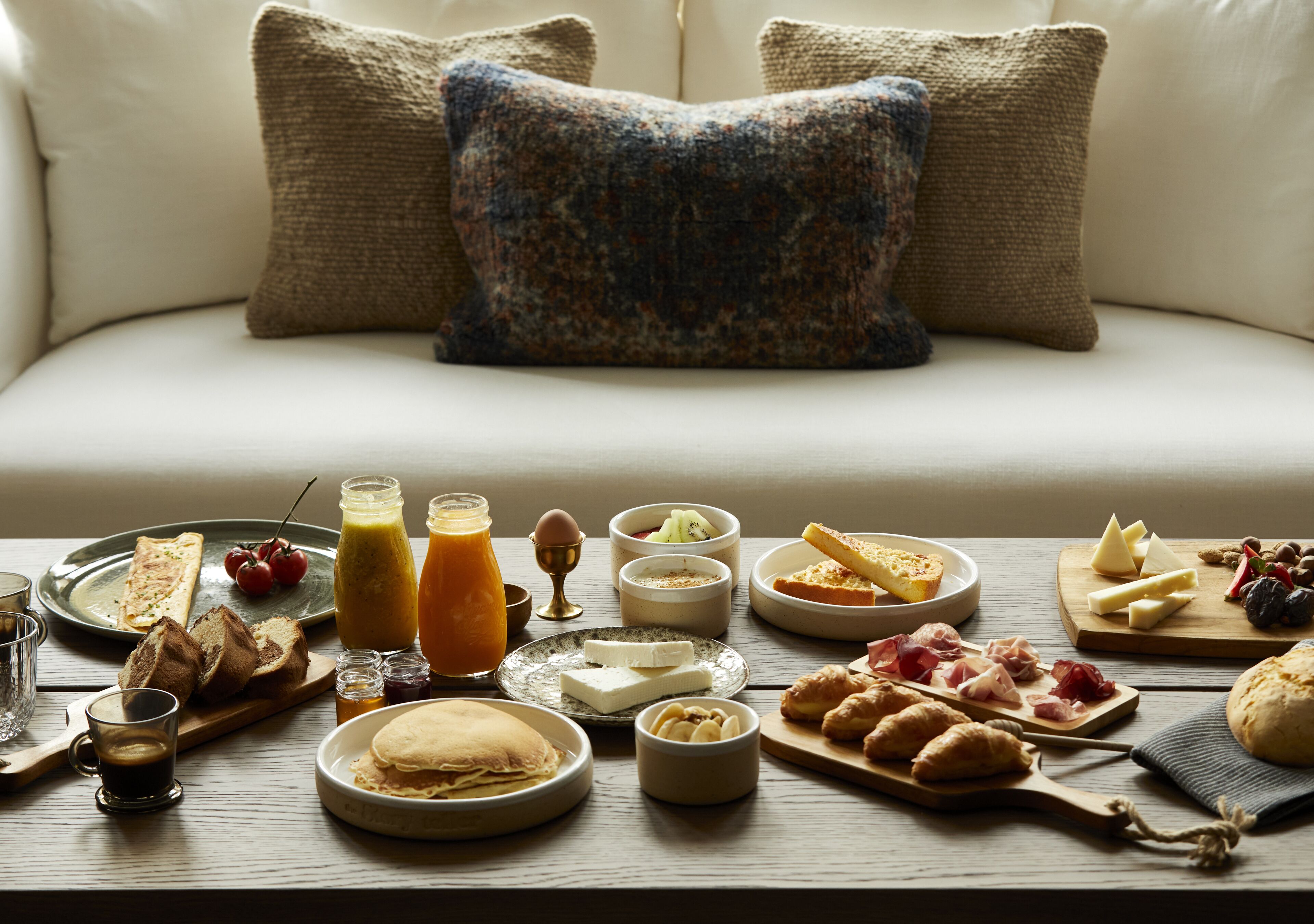 A breakfast table with various foods and drinks, a sofa with pillows, and a wooden table.