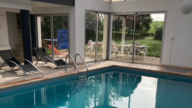 Una piscina interior con paredes de vidrio transparente, suelo de baldosas y algunas sillas de descanso.