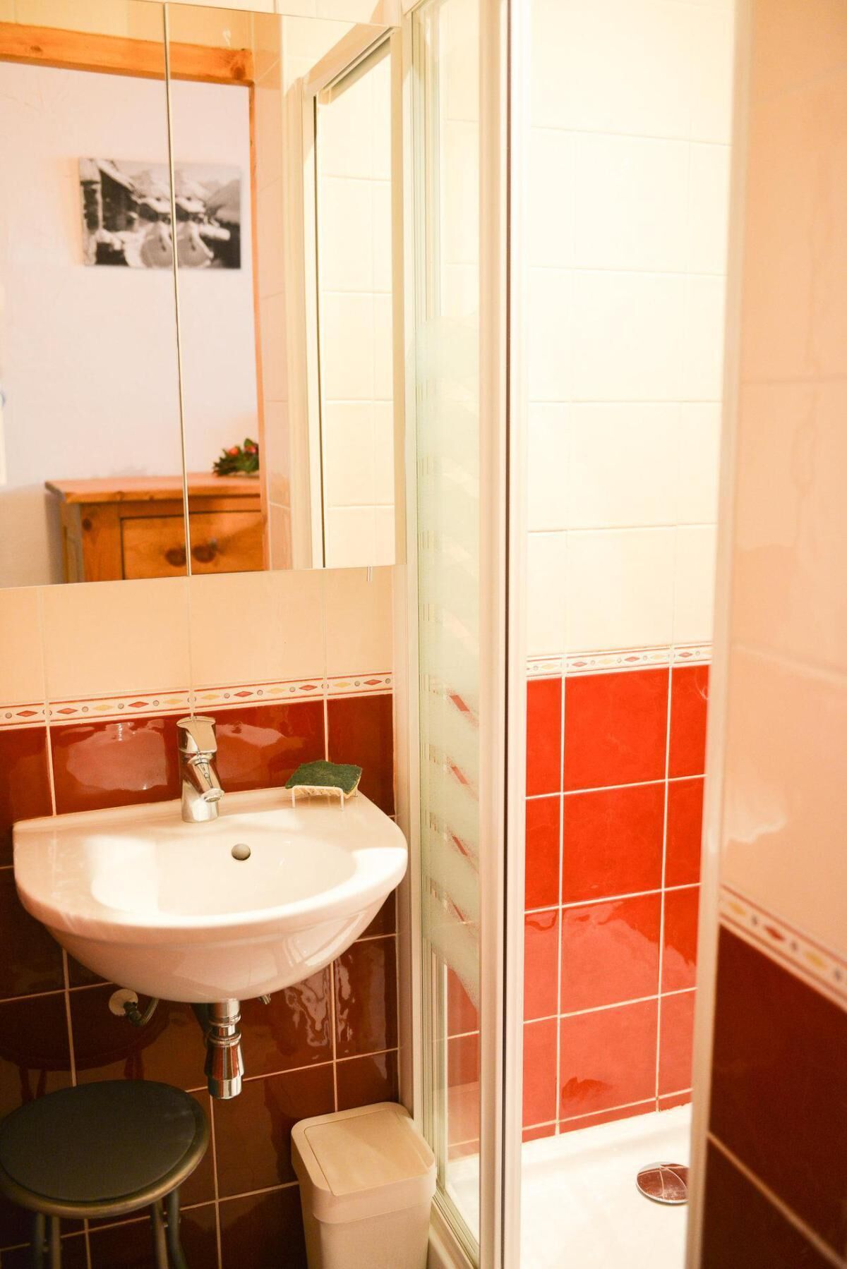 Un bagno con un lavandino bianco, uno sgabello, un cestino e uno spazio doccia con piastrelle rosse.