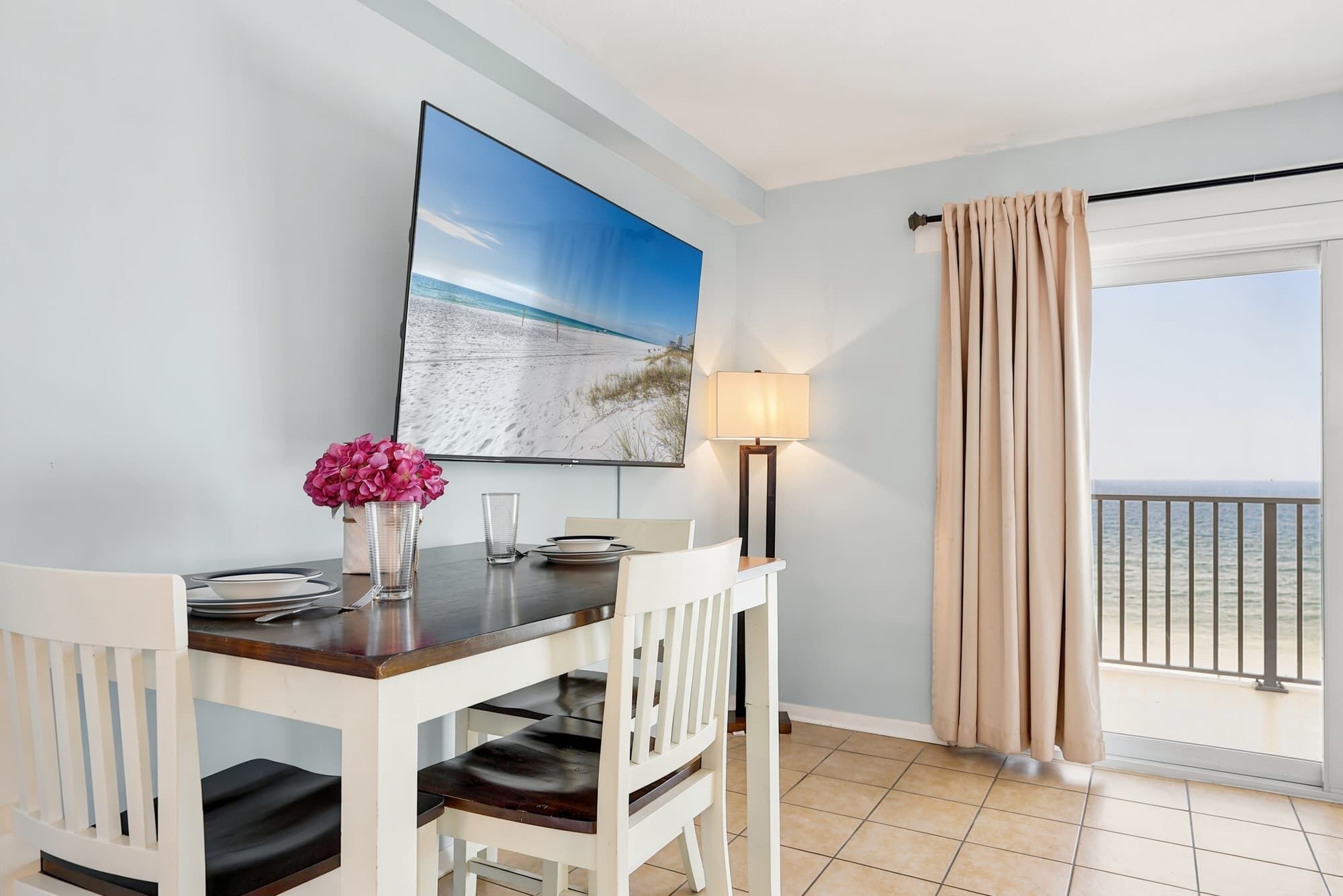 A dining area with a table, chairs, a painting, a lamp, and a view of the sea through a window.