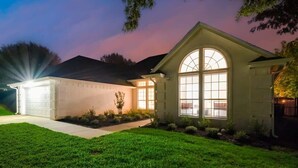 Uma casa residencial com garagem bem iluminada, uma grande janela em arco e um jardim frontal bem cuidado.