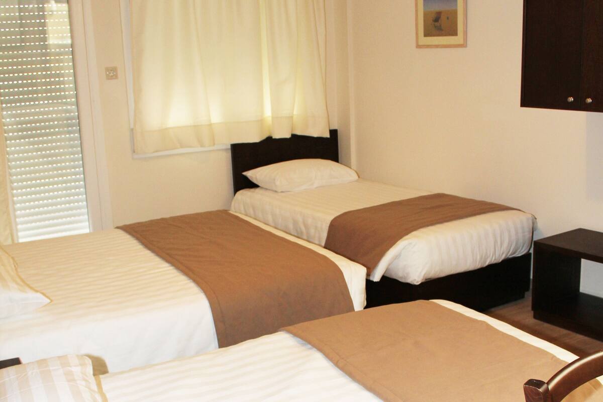 A hotel room with two single beds, a wooden chair, a small table, and a wall-mounted air conditioner.