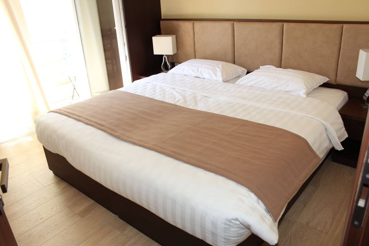A hotel room with a large bed, a bedside table with a lamp, and a view of the outside.