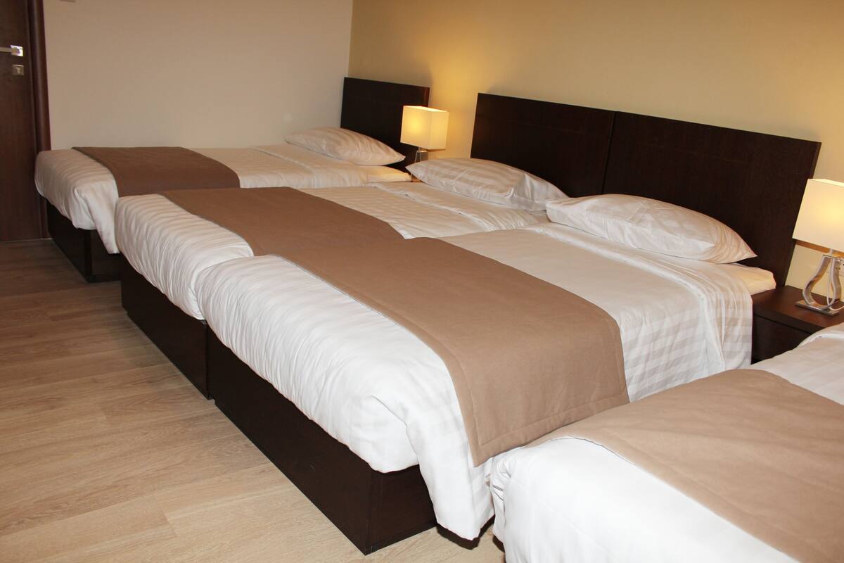 A hotel room with three single beds, each with a brown blanket and white linens. There are bedside lamps on each nightstand.