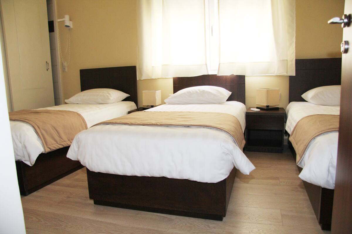 A hotel room with three single beds, wooden flooring, and a neutral color scheme.