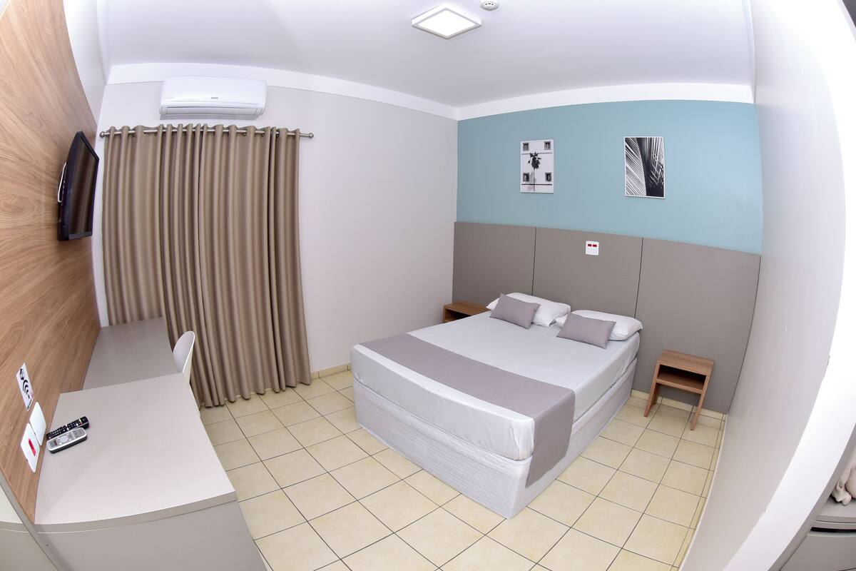 Habitación de hotel con cama, mesitas de noche, un escritorio con silla, unidad de aire acondicionado, cortinas y una obra de arte colgada en la pared.