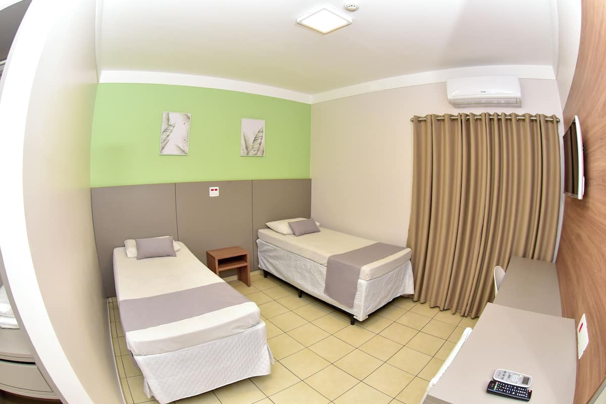 Habitación de hotel con dos camas individuales, una mesita pequeña, un escritorio con un control remoto y una ventana con cortinas.