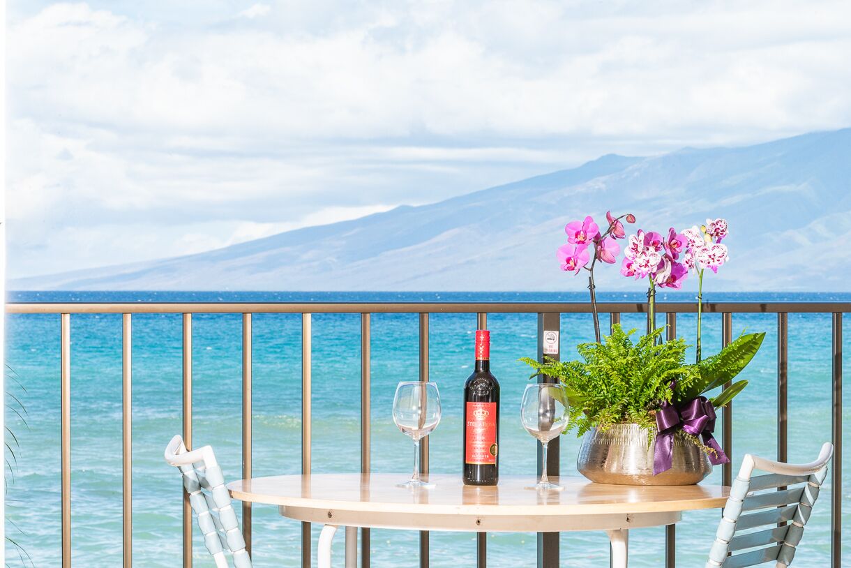Ett bord med en vinflaska, två glas och en krukväxt med blommor på en balkong med utsikt över en sjö eller ett hav och berg.