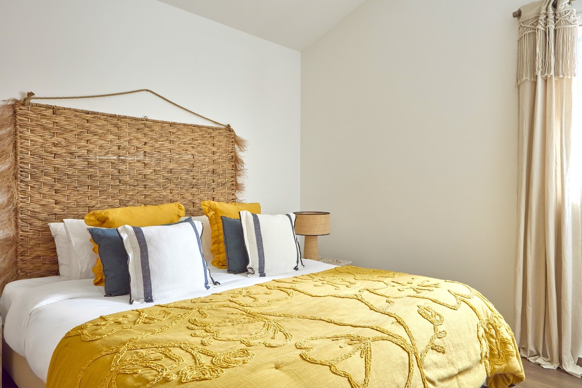 A bed with a woven headboard, multiple pillows in various colors, a yellow bedspread, and a bedside lamp.