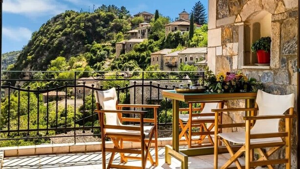 Un balcón con sillas y mesa de madera, con vistas a una colina con edificios y follaje.