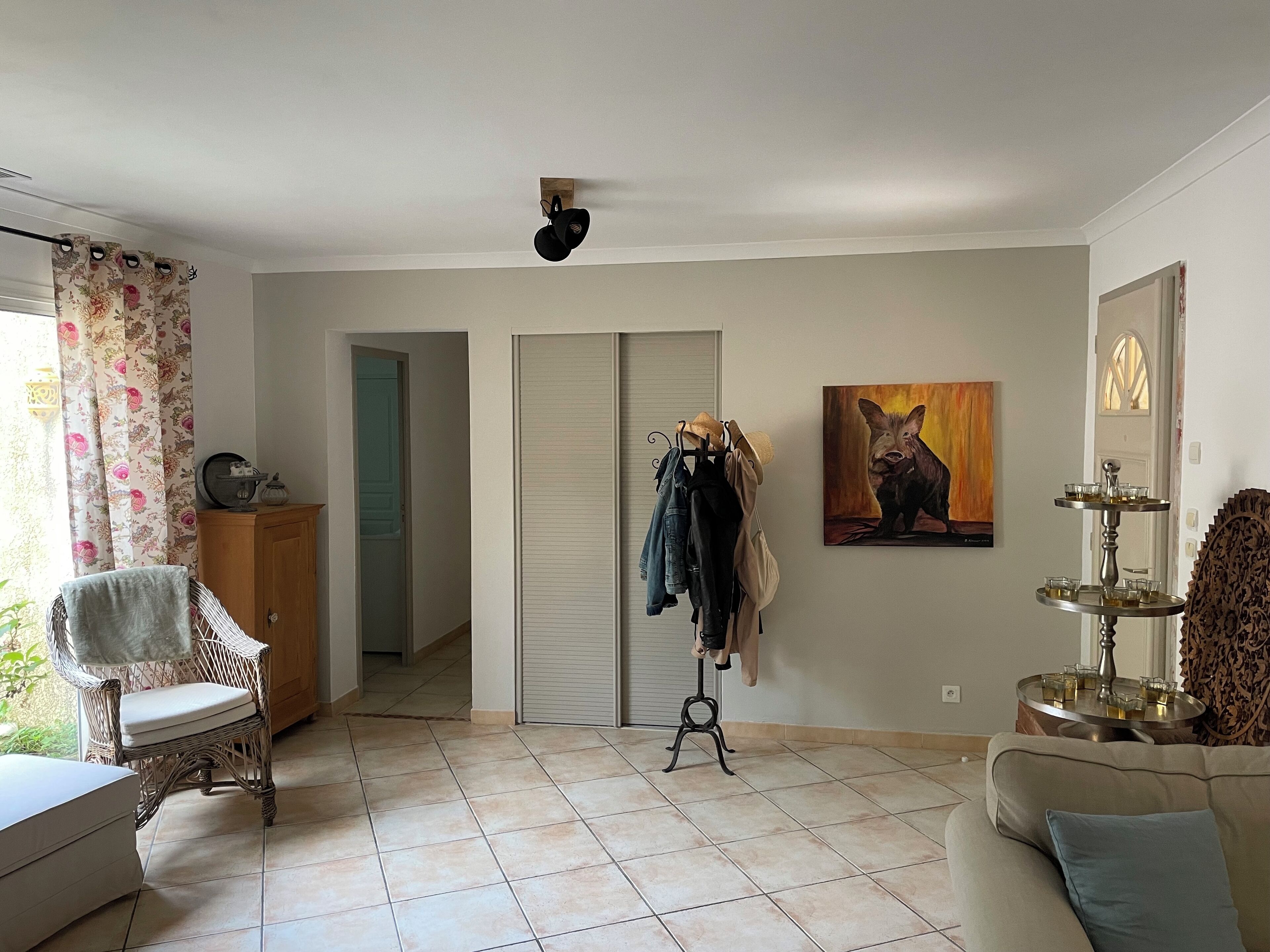 Une pièce comprenant un fauteuil en osier, un canapé, un porte-manteau, une armoire et un tableau représentant un sanglier.