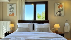 A neatly made bed with white bedding and two pillows, a window with a view of palm trees, and two bedside lamps on wooden nightstands. - Villa Metapothia in Gavalohori (Apokoronas)