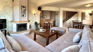A spacious living room with a stone fireplace, a large sofa, and a dining area with a staircase. - Villa Metapothia in Gavalohori (Apokoronas)