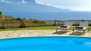 A pool with two lounge chairs, a wooden fence, and a view of the sea and mountains. - Villa Afroditi in Spartia (Kefalonia)