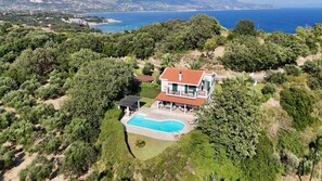 A two-story house with a swimming pool, surrounded by dense greenery and overlooking a coastal area.