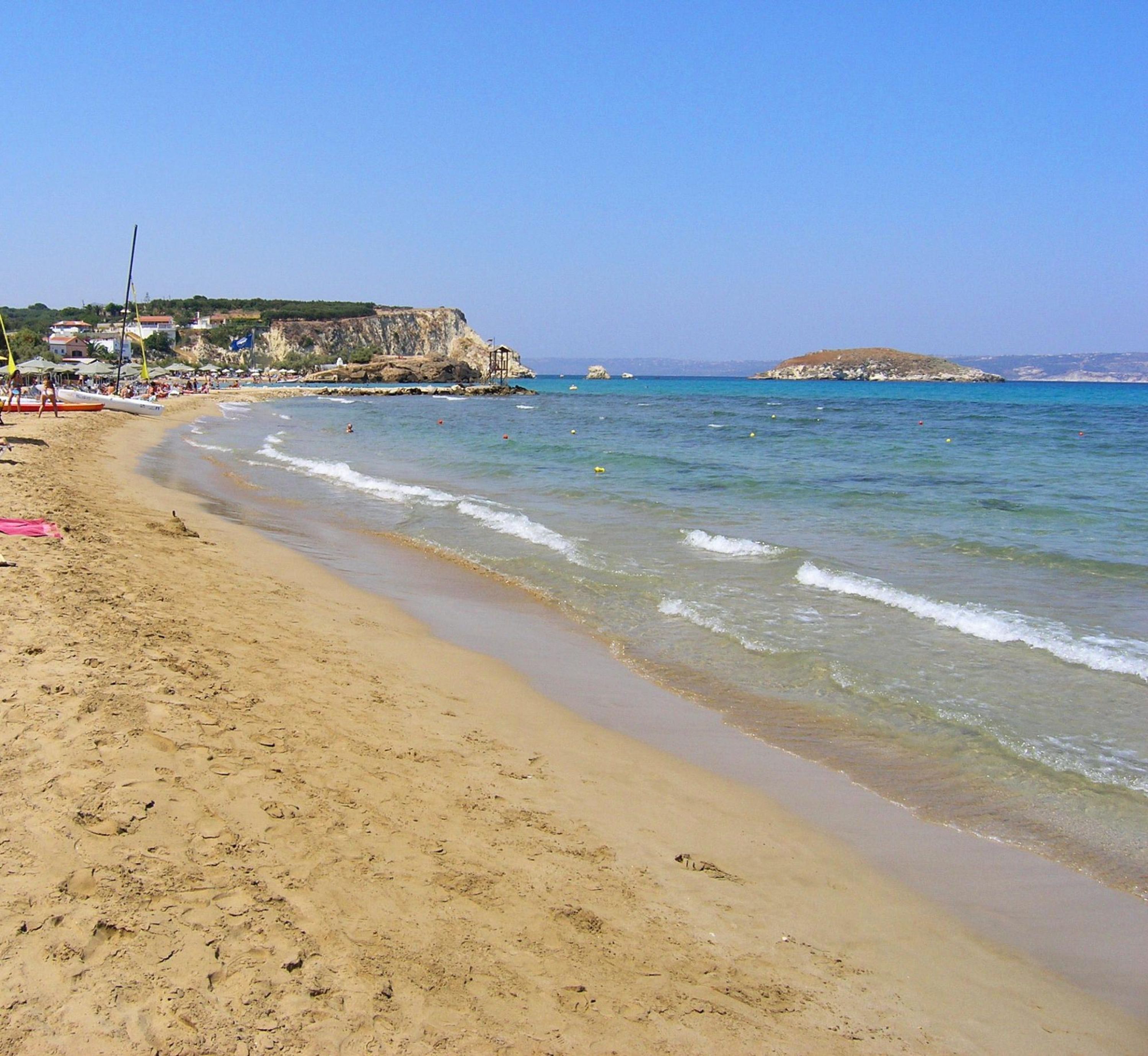 Ein sandiger Strand mit klarem, blauem Wasser, ein felsiger Felsen im Hintergrund und einige Gebäude am Ufer.