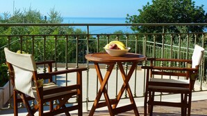 Un balcon avec une table en bois et deux chaises, un panier de fruits et une vue sur la mer.