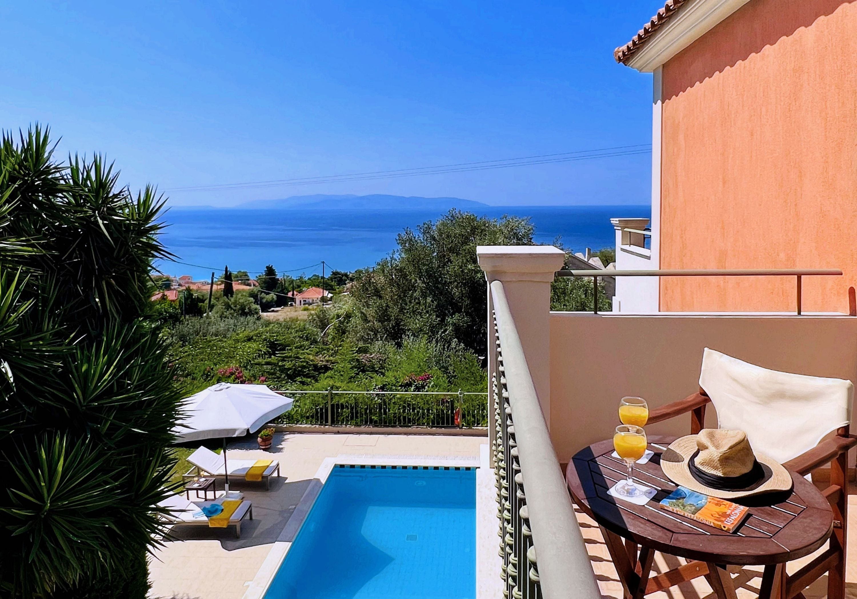 Blick über den Pool mit freier Sicht auf das Meer, eine Lounge-Ecke mit Tisch und Stühlen sowie ein Gebäude mit Balkon.