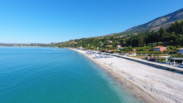 Une plage côtière aux eaux turquoise et limpides, bordée d’un rivage sablonneux, avec des collines en arrière-plan parsemées de bâtiments.
