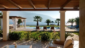 Ein überdachter Außenbereich mit Pool, Liegestühlen und Blick auf das Meer und die Berge.