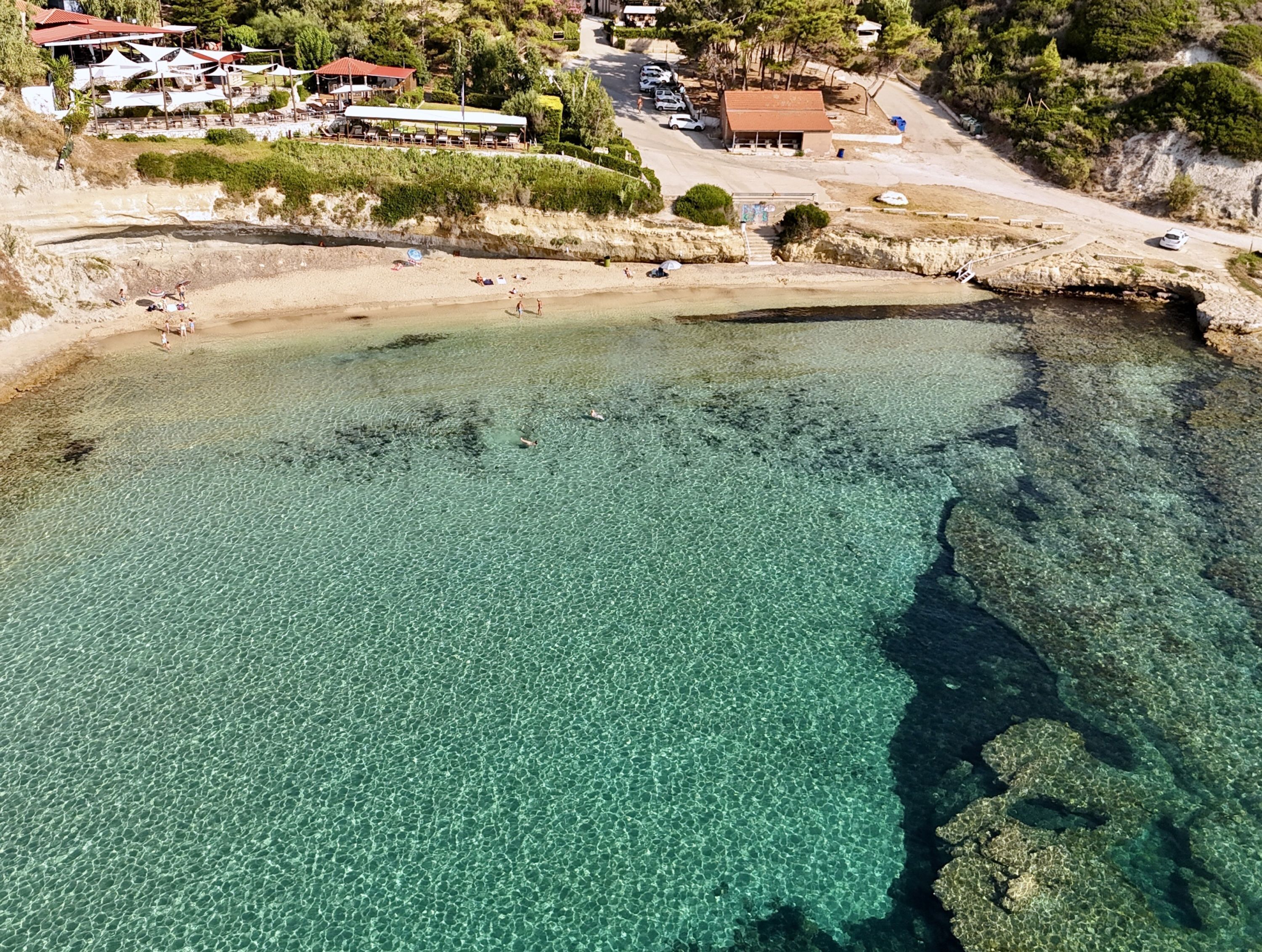 A coastal area with clear shallow waters, a sandy beach, and a few buildings in the background.