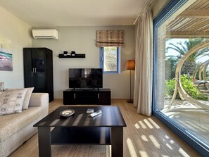 A modern living room with a flat-screen TV on a black stand, a sofa with patterned cushions, a coffee table, and a large window with curtains. - Villa Odysseus in Spartia (Kefalonia)