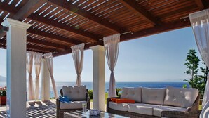 An outdoor seating area with wicker furniture, a glass-top table, and a view of the sea. - Villa Odysseus in Spartia (Kefalonia)