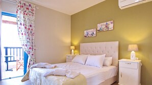 A bedroom with a large bed, a nightstand with a lamp, a small cabinet, and two wall art pieces. - Villa Odysseus in Spartia (Kefalonia)