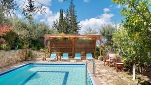 Una piscina con agua azul cristalina, rodeada de baldosas de piedra. Se observan sillones de descanso y una pérgola de madera con celosía trenzada.