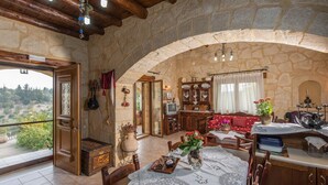 Una habitación rústica con paredes de piedra, techo de madera, un gran arco de entrada, una mesa de comedor con mantel de encaje y un televisor.