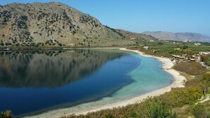 Paisaje costero con un lago tranquilo, una playa arenosa y montañas al fondo.