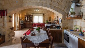 Un comedor rústico con paredes de piedra, una mesa de madera dispuesta para cuatro comensales, y a sus lados una cocina con estufa y horno, así como una zona de estar acogedora con un sofá rojo.