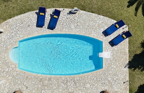 An oval-shaped swimming pool with a tiled edge, surrounded by a stone patio with lounge chairs and a small white structure.
