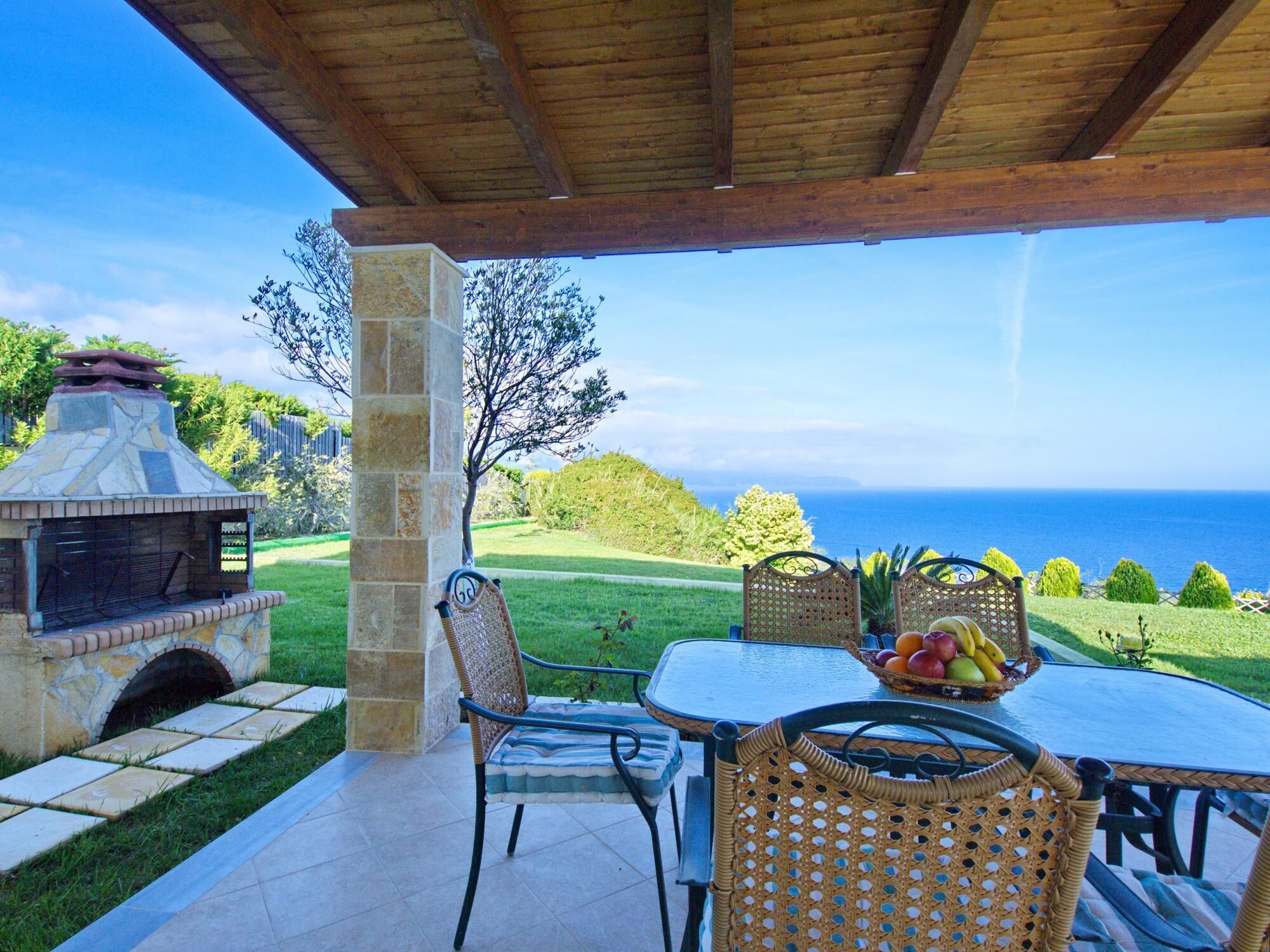 An outdoor patio with a stone barbecue, wicker chairs, a table with fruit, and a view of the ocean.