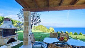 Une terrasse extérieure avec un barbecue en pierre, des chaises en osier, une table garnie de fruits et une vue sur l’océan.