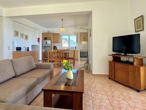 A living room with a sofa, a coffee table with a vase, a television on a wooden stand, and a kitchen in the background.