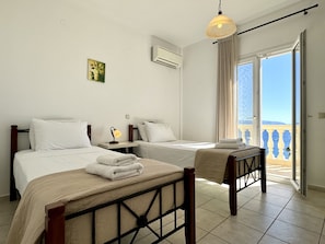 A hotel room with two beds, a bedside table with a lamp, a window with curtains, and a view of the sea.