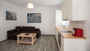Un espacio habitable compacto con un sofá, una mesa de centro, una zona de cocina con un lavabo y una cafetera roja, y dos cuadros colgados en la pared.