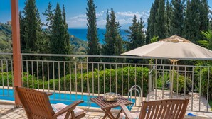 A balcony with a pool, wooden chairs, and an umbrella. - Villa Mounda Poseidon (Kefalonia)