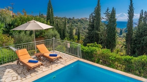 A poolside area with lounge chairs and an umbrella, overlooking a scenic landscape with trees and a view of the sea. - Villa Mounda Poseidon (Kefalonia)