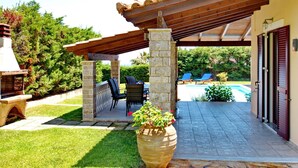 Eine überdachte Terrasse mit Steinpfeilern, Gartenmöbeln, einem Swimmingpool und einem Grillbereich aus Stein.