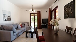 Ein Wohnzimmer mit einer Couch, einem Couchtisch, einem Esstisch, Stühlen, einem Fernseher und einer Vase mit Blumen.