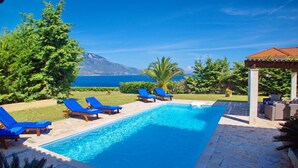 Ein Swimmingpool mit blauen Liegestühlen, einer Steindeck und Blick auf das Meer und die Berge.