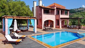 A pool area with lounge chairs and a swing chair, a covered seating area, and a building with a red-tiled roof and stone chimneys.