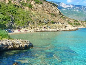 A coastal beach with clear turquoise water, rocky cliffs, and sparse vegetation.