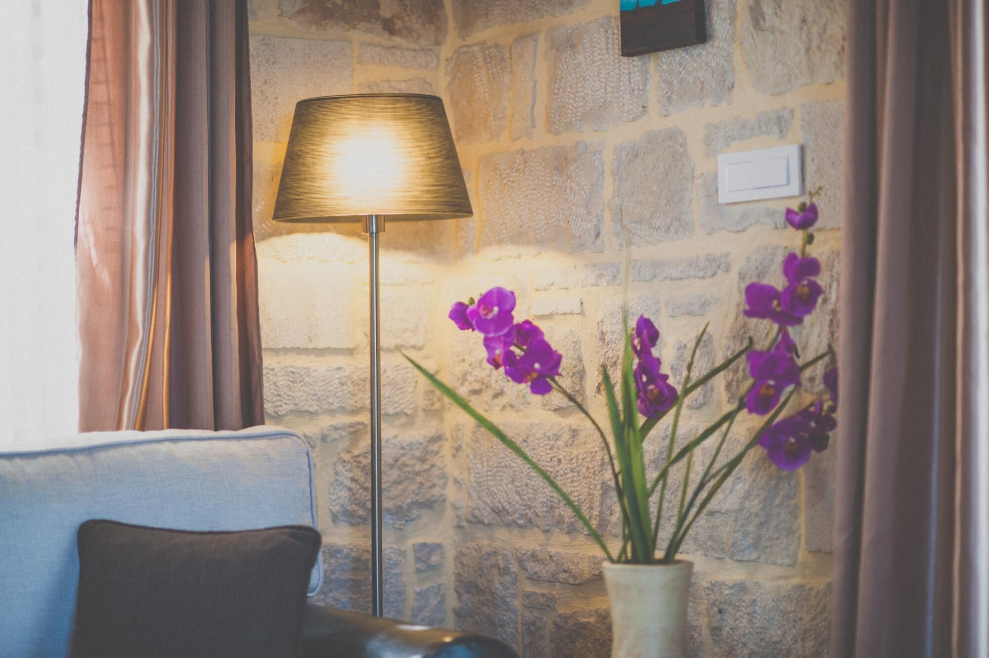 A room with a stone wall, a floor lamp, and a vase with purple flowers.